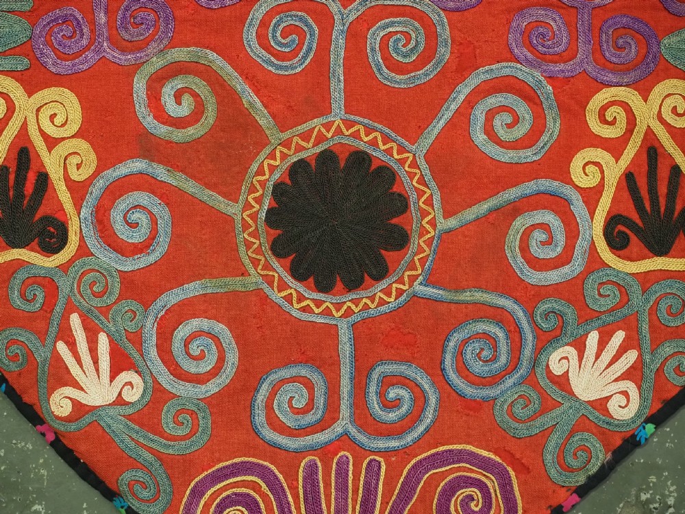 A shield-shaped red Central Asian textile featuring dense silk embroidery with swirling scroll motifs and floral rosettes. View VII