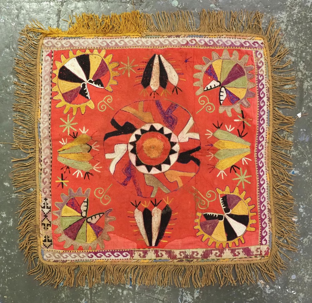 A square red embroidered textile featuring bold central circular motifs, tribal geometric shapes, and a fringed outer border. View II