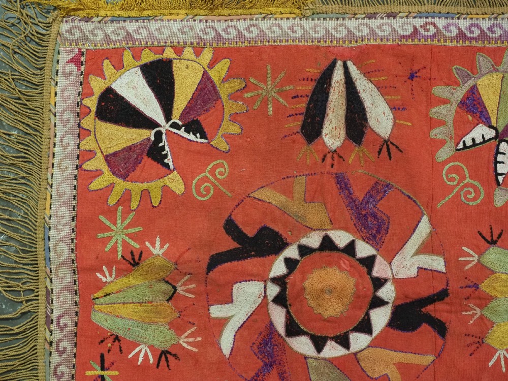 A square red embroidered textile featuring bold central circular motifs, tribal geometric shapes, and a fringed outer border. View III