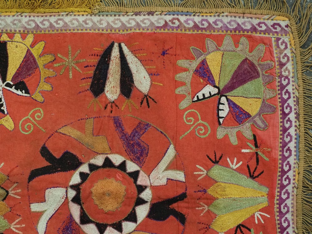 A square red embroidered textile featuring bold central circular motifs, tribal geometric shapes, and a fringed outer border. View IV
