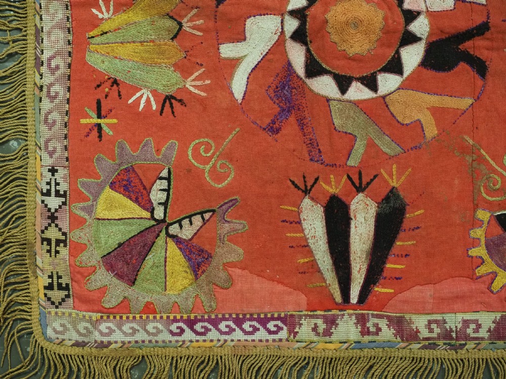 A square red embroidered textile featuring bold central circular motifs, tribal geometric shapes, and a fringed outer border. View V