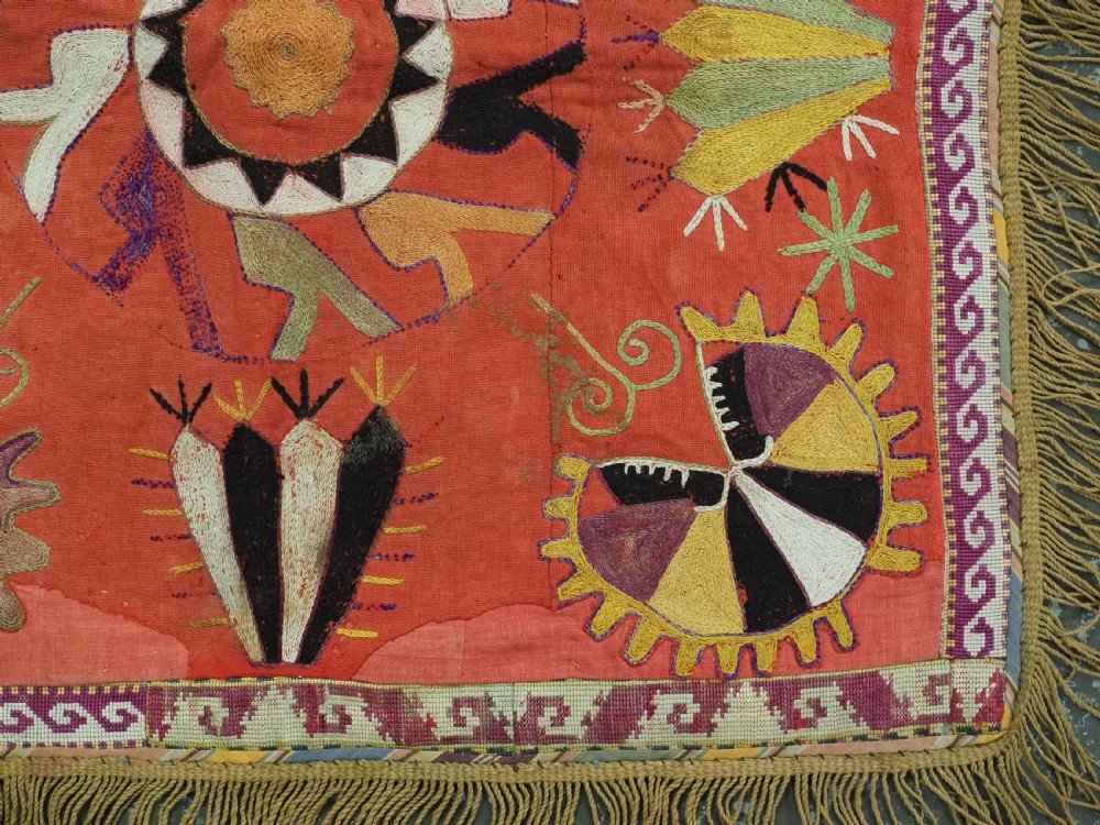 A square red embroidered textile featuring bold central circular motifs, tribal geometric shapes, and a fringed outer border. View VI