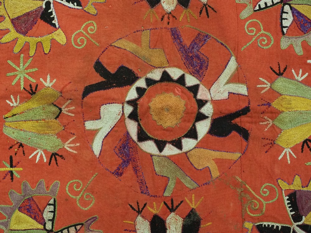 A square red embroidered textile featuring bold central circular motifs, tribal geometric shapes, and a fringed outer border. View VII