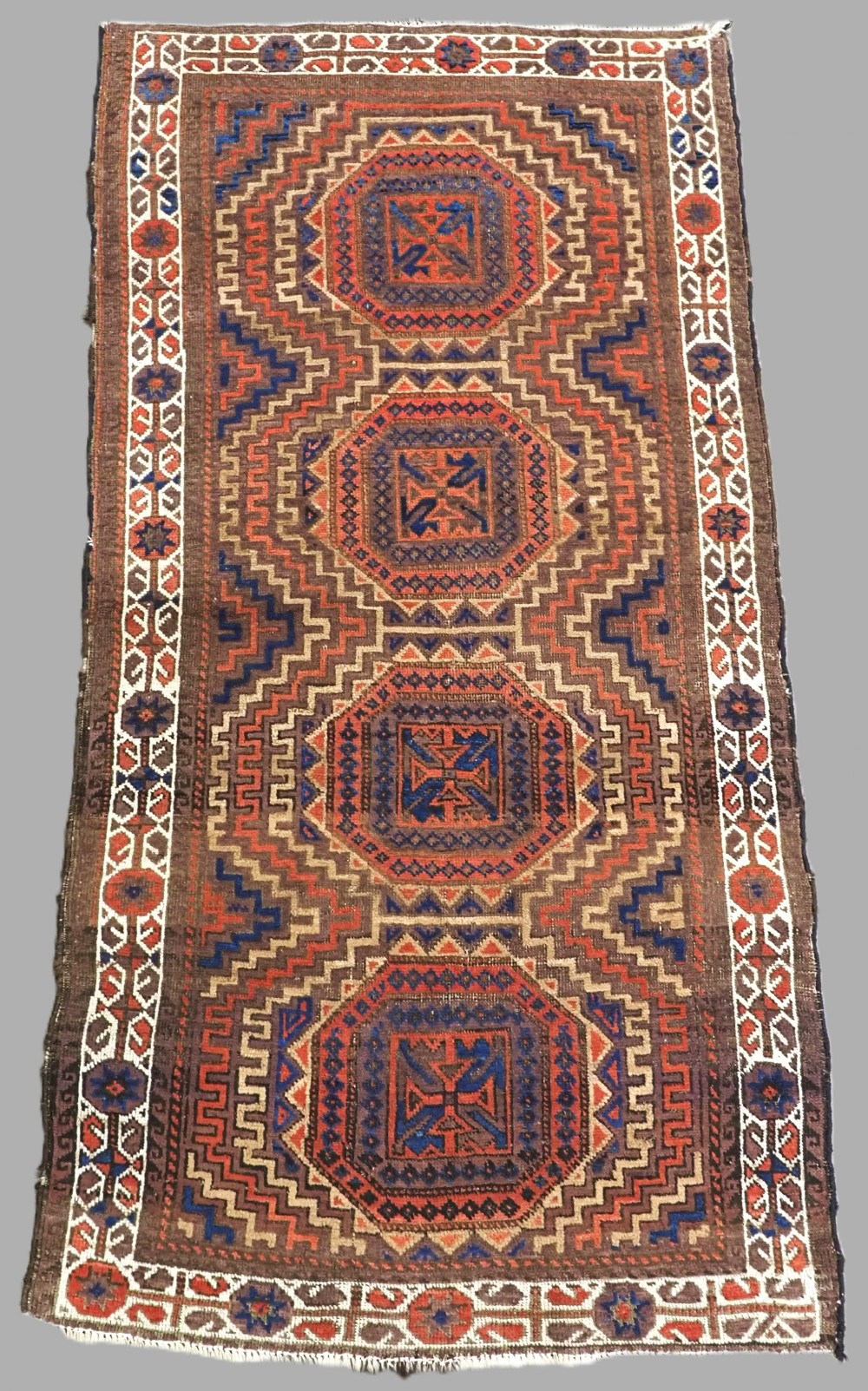 An antique hand-knotted wool rug featuring four central octagonal guls framed by stepped geometric borders in dark brown, terracotta, blue, and cream hues.