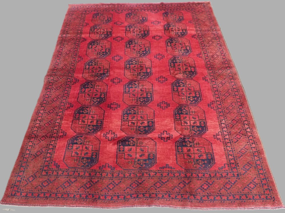 A hand-knotted antique Afghan wool carpet in warm red featuring dark blue repeating octagonal gul motifs and geometric borders.