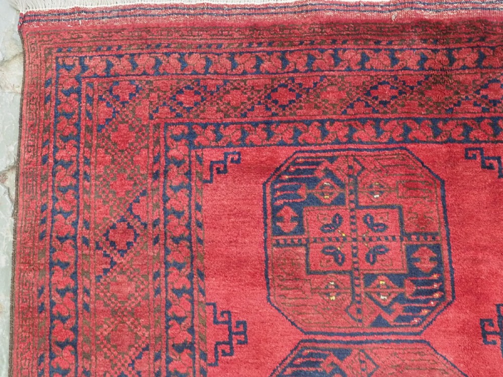 A hand-knotted antique Afghan wool carpet in warm red featuring dark blue repeating octagonal gul motifs and geometric borders. View VI