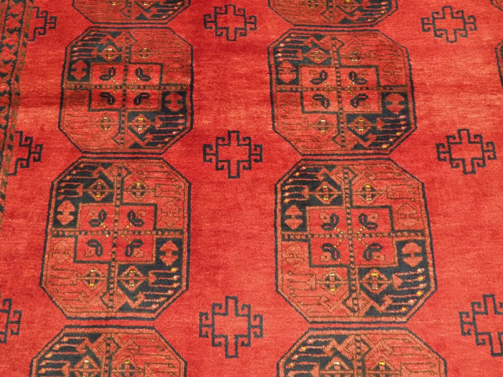 A hand-knotted antique Afghan wool carpet in warm red featuring dark blue repeating octagonal gul motifs and geometric borders. View IX