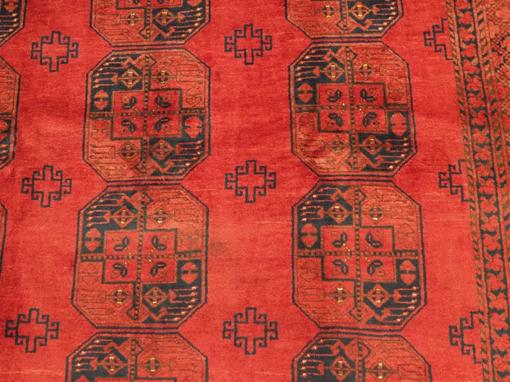 A hand-knotted antique Afghan wool carpet in warm red featuring dark blue repeating octagonal gul motifs and geometric borders. View X