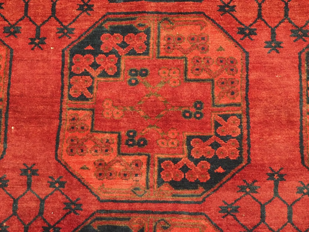 An antique hand-knotted Afghan Ersari wool carpet featuring dark blue octagonal gul motifs arranged in rows on a warm red field. View IX
