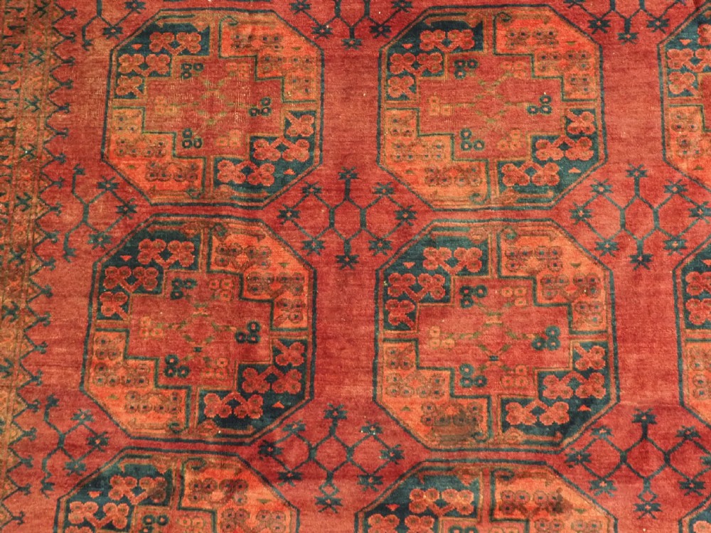 An antique hand-knotted Afghan Ersari wool carpet featuring dark blue octagonal gul motifs arranged in rows on a warm red field. View X