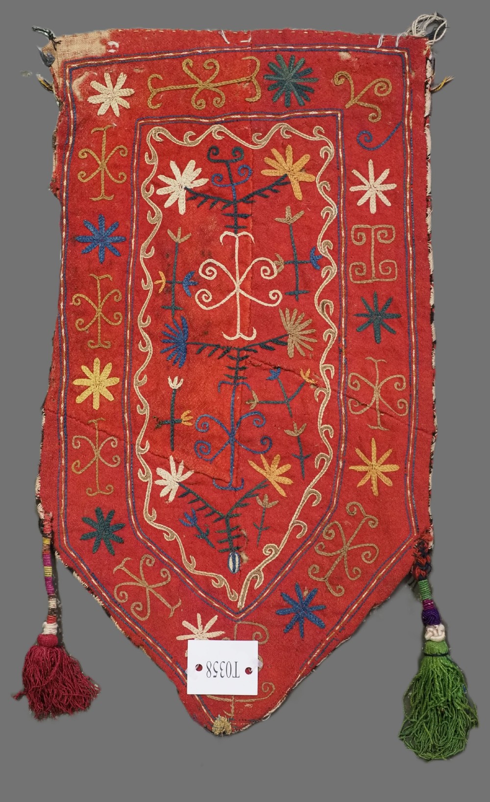 A vibrant red wool embroidered tribal textile bag featuring intricate polychrome scroll motifs in yellow, blue, purple, and green, finished with tassels at the bottom corners. View II