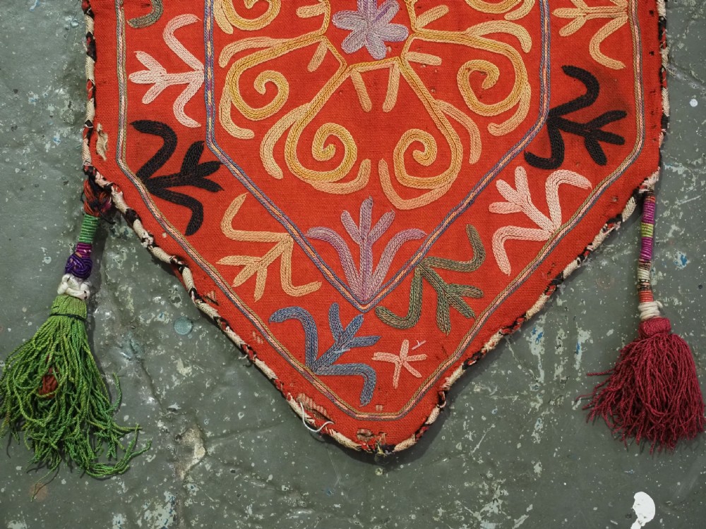 A vibrant red wool embroidered tribal textile bag featuring intricate polychrome scroll motifs in yellow, blue, purple, and green, finished with tassels at the bottom corners. View V