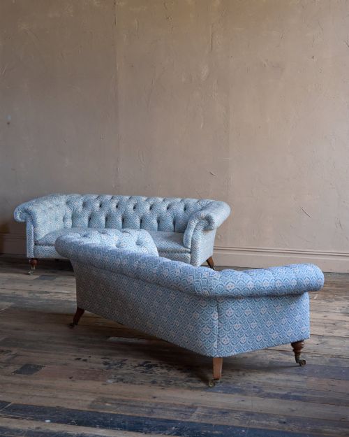 fig 3. A pair of antique serpentine Chesterfield sofas featuring light blue patterned fabric, deep button-tufted backrests, rolled arms, and turned wooden legs on brass castors.
