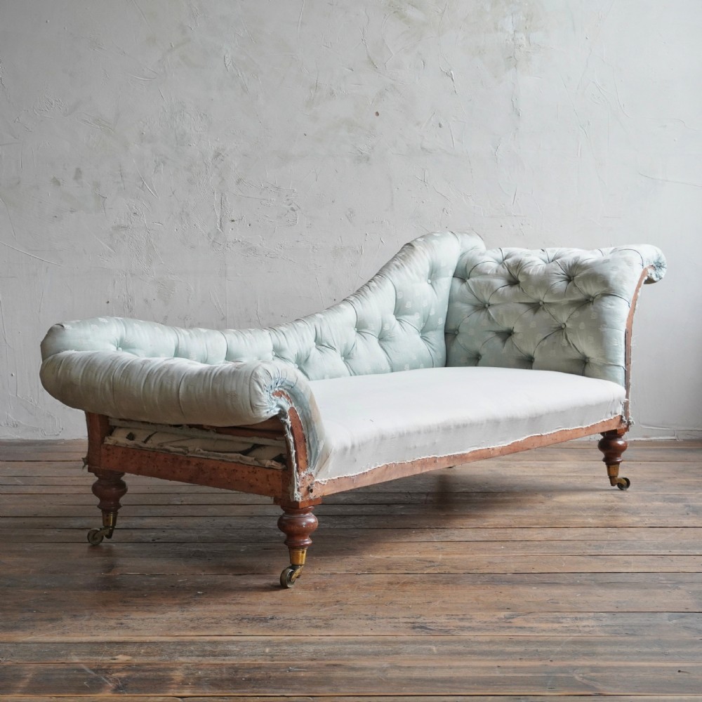 An early Victorian upholstered chaise longue featuring light blue deep-buttoned fabric, a scrolled backrest, and turned wood legs with brass castors.