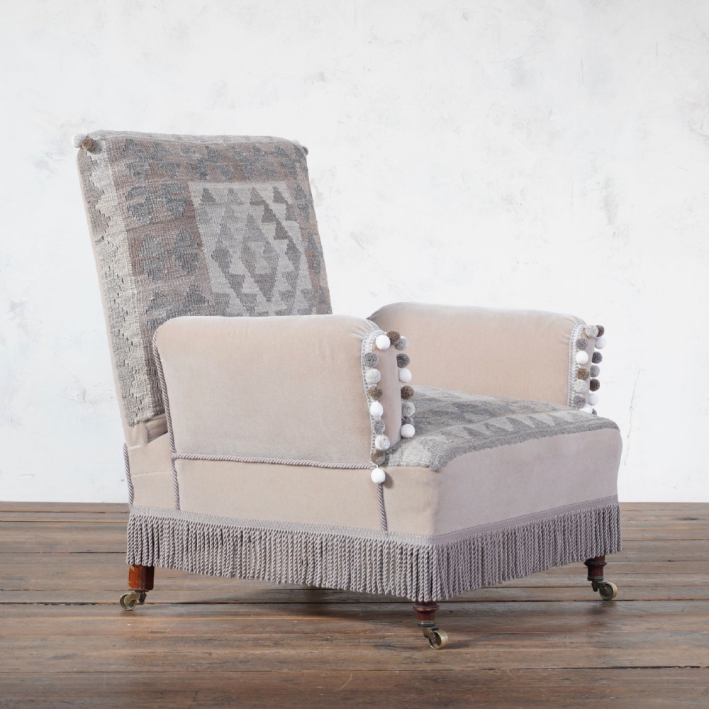 A Victorian style armchair upholstered with grey geometric kilim textile on the backrest and seat, trimmed with beige velvet, pom-poms, fringe, and mounted on turned legs with brass castors.