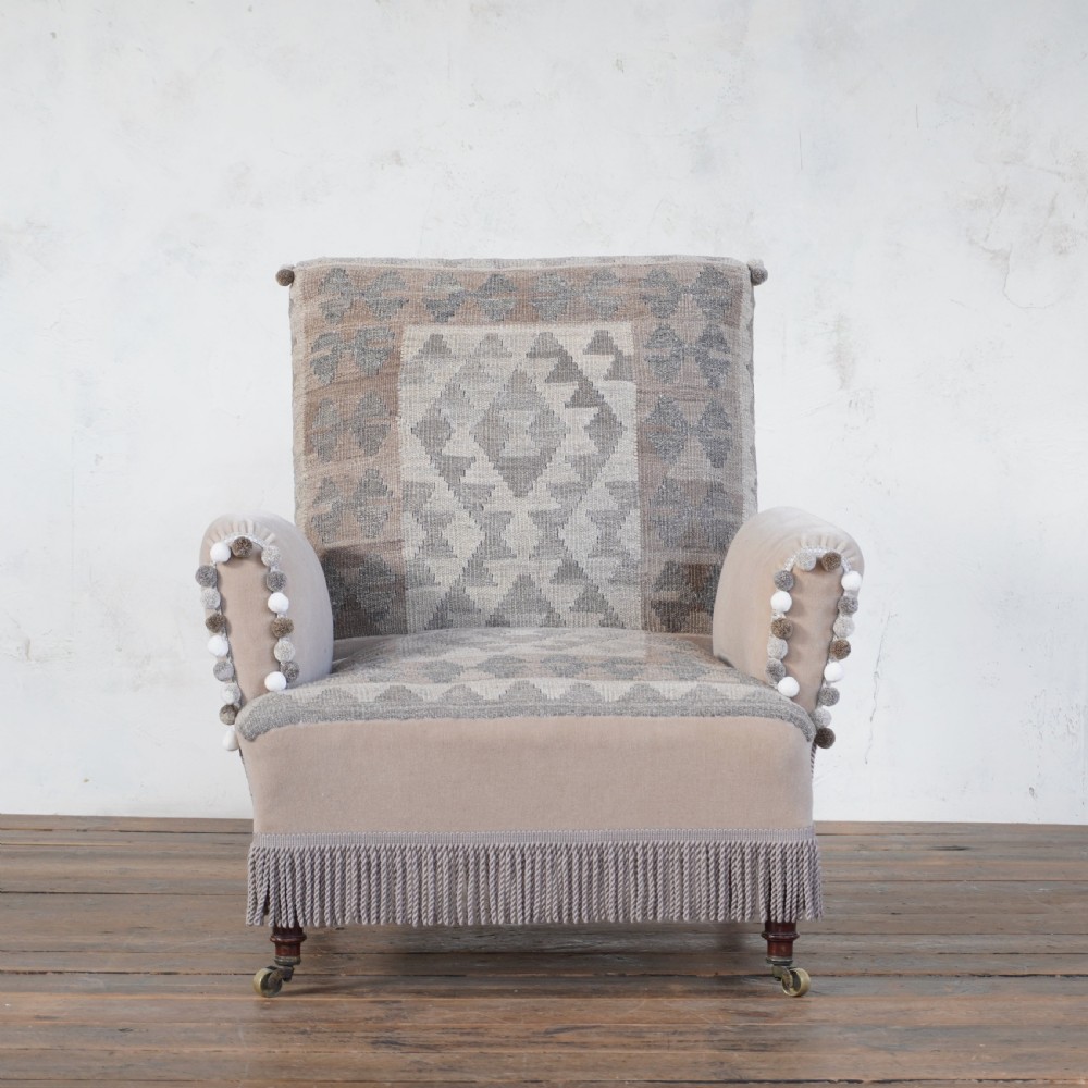 A Victorian style armchair upholstered with grey geometric kilim textile on the backrest and seat, trimmed with beige velvet, pom-poms, fringe, and mounted on turned legs with brass castors. View II