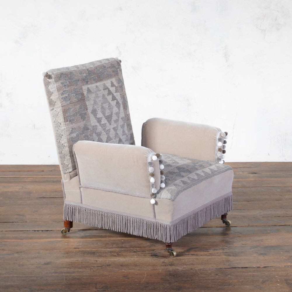 A Victorian style armchair upholstered with grey geometric kilim textile on the backrest and seat, trimmed with beige velvet, pom-poms, fringe, and mounted on turned legs with brass castors. View III