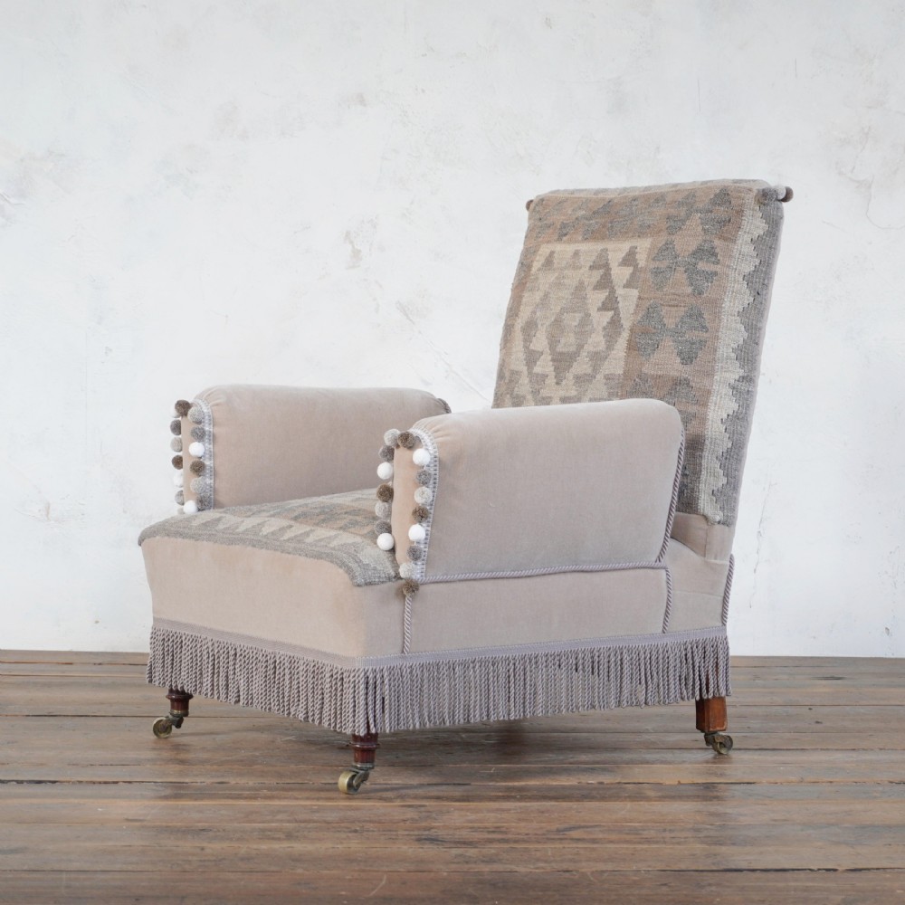 A Victorian style armchair upholstered with grey geometric kilim textile on the backrest and seat, trimmed with beige velvet, pom-poms, fringe, and mounted on turned legs with brass castors. View V