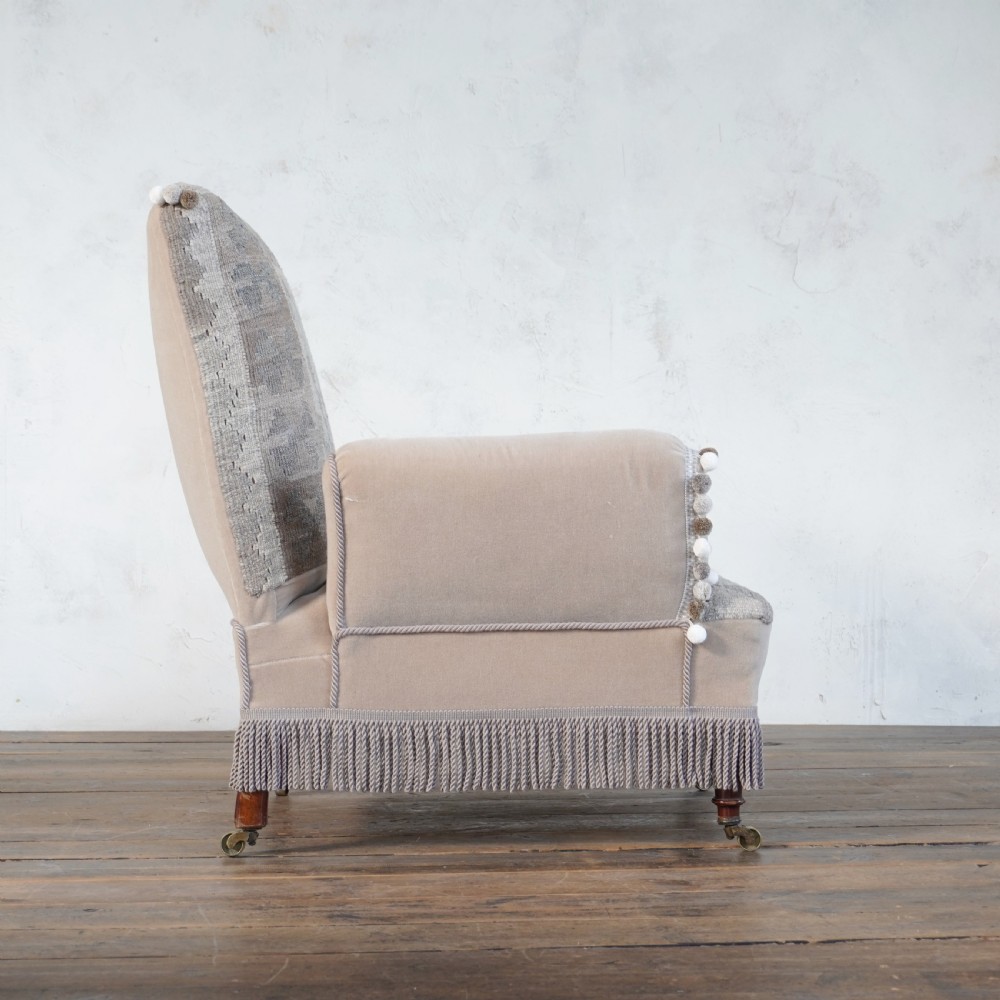 A Victorian style armchair upholstered with grey geometric kilim textile on the backrest and seat, trimmed with beige velvet, pom-poms, fringe, and mounted on turned legs with brass castors. View VI