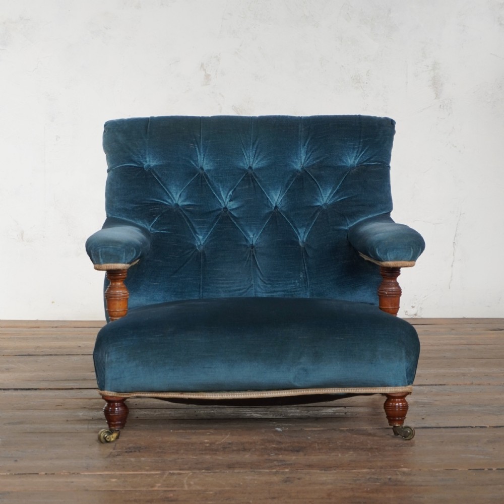 An antique Howard and Sons open armchair upholstered in dark blue velvet, featuring button-tufting on the backrest and turned wooden legs with brass castors. View VI
