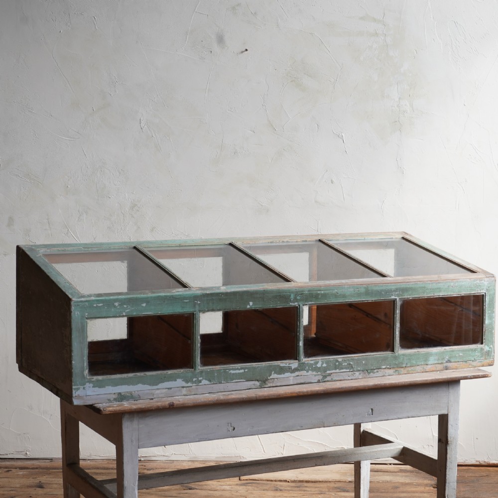 An antique sloped countertop display cabinet featuring four glazed compartments and a distressed green painted timber frame.