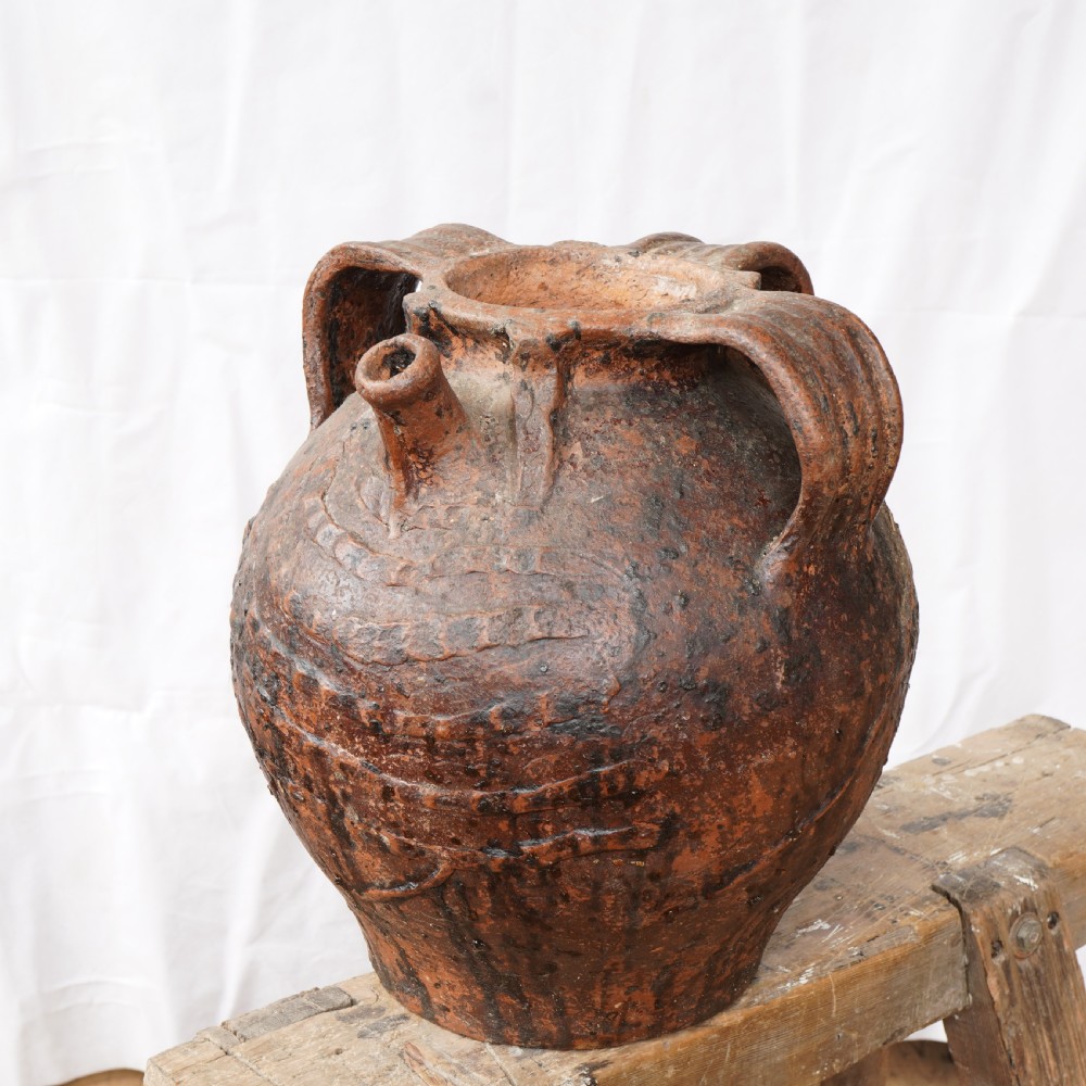 A rustic brown terracotta oil jug featuring a small spout, sturdy side handles, and incised horizontal banding on its bulbous body.