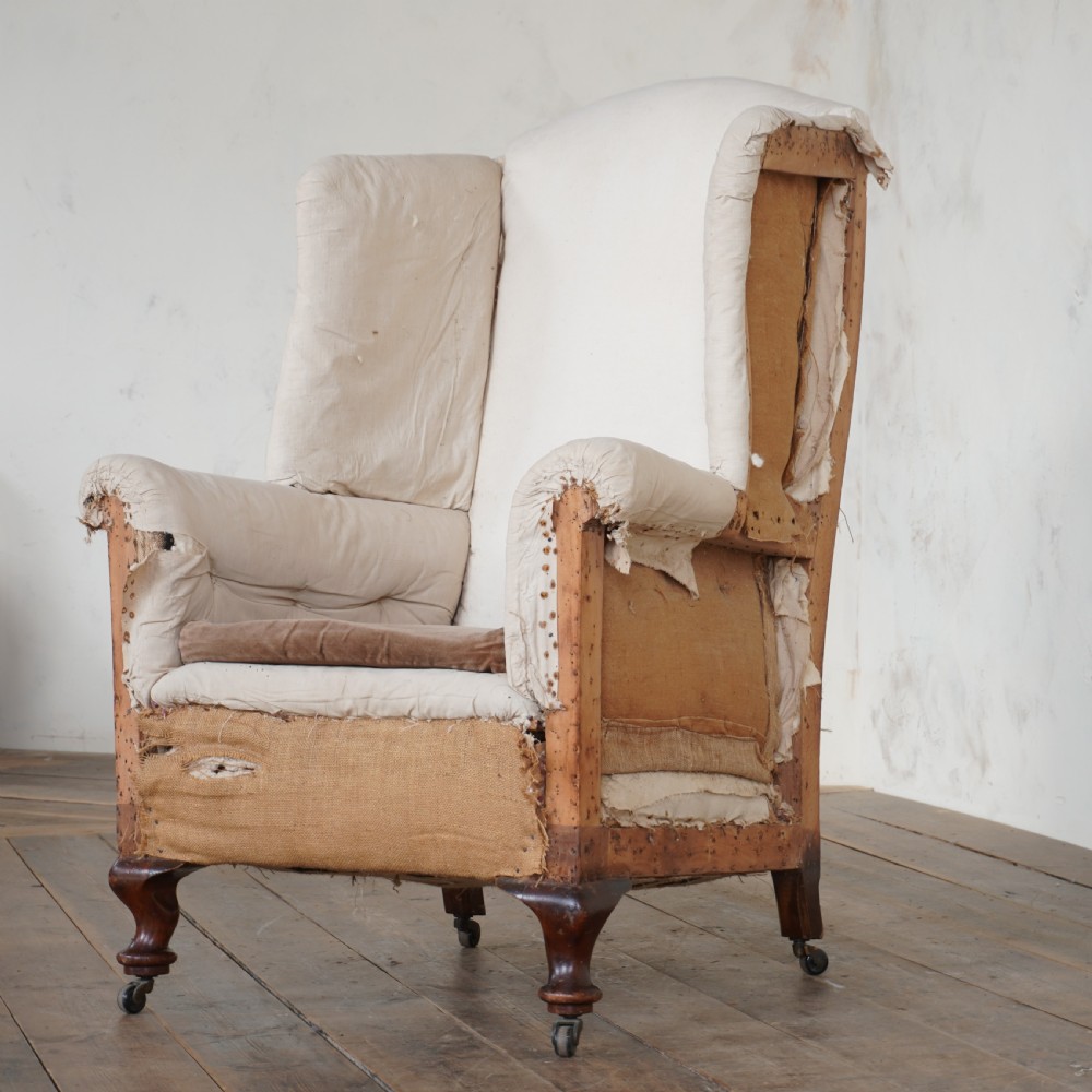 An antique wingback armchair shown in a stripped-down state with exposed wooden frame, hessian webbing, and calico under-upholstery.