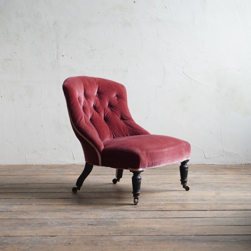 A low Victorian slipper chair upholstered in deep buttoned rose-pink velvet with turned dark front legs on brass castors.