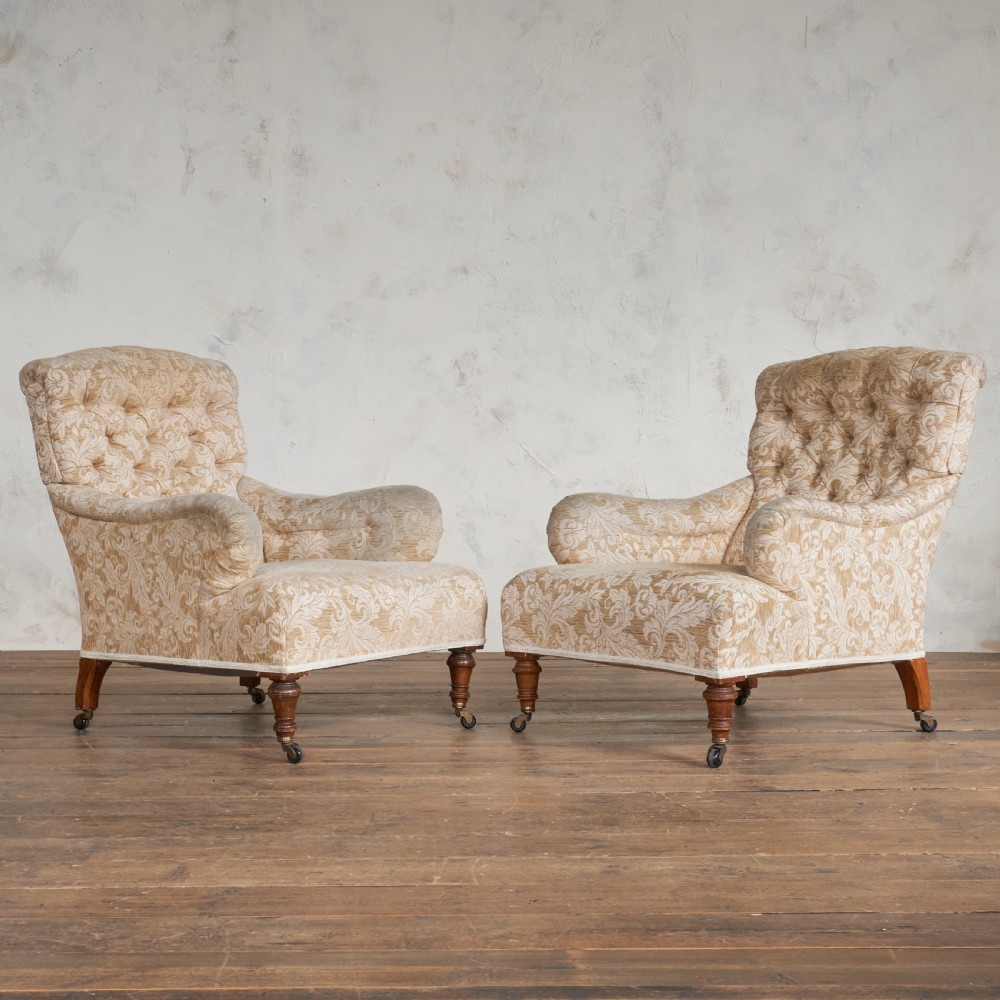 A pair of Victorian Howard style armchairs upholstered in a patterned beige fabric, featuring buttoned backs and turned wooden legs with brass castors.