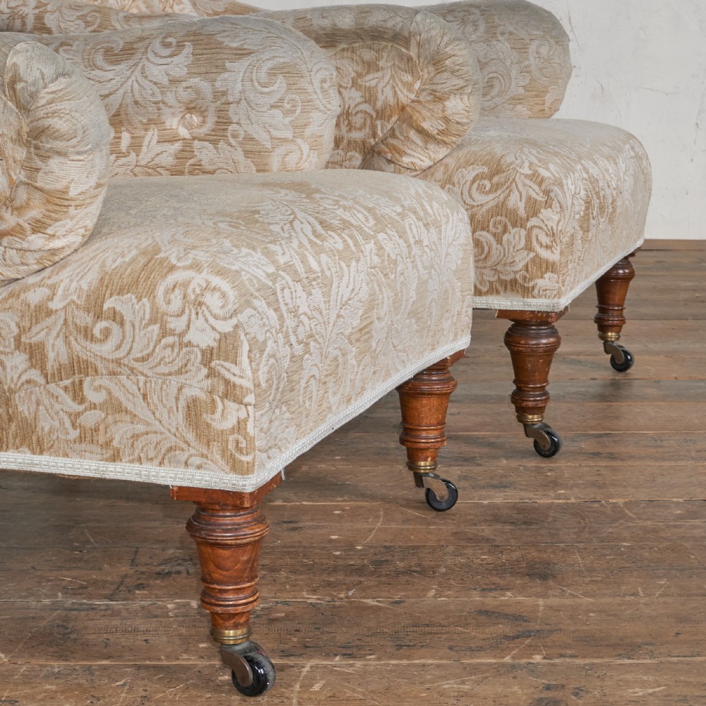 A pair of Victorian Howard style armchairs upholstered in a patterned beige fabric, featuring buttoned backs and turned wooden legs with brass castors. View V