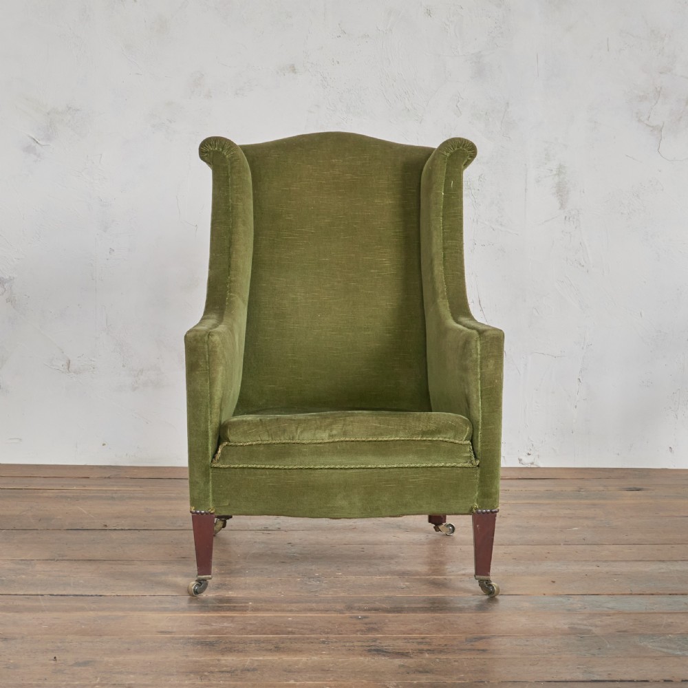 An antique green velvet wingback armchair featuring rolled arms, dark wooden legs, and brass castors. View II