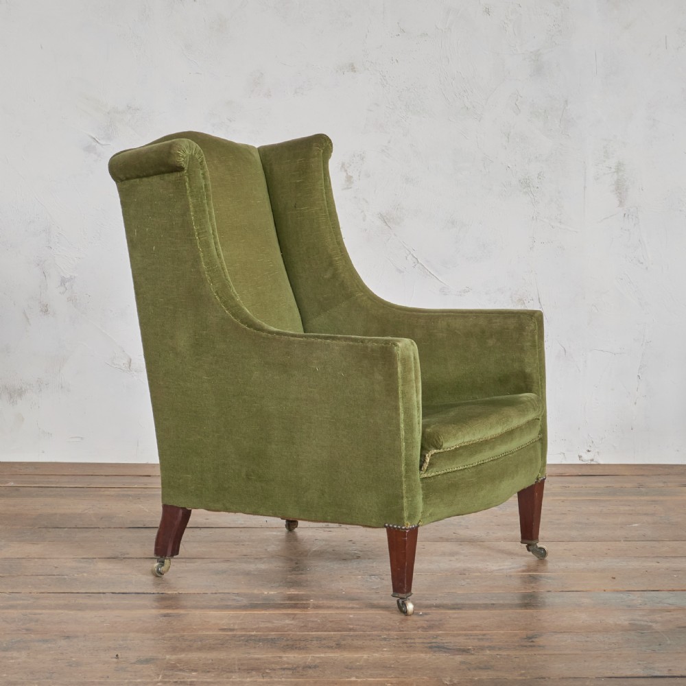 An antique green velvet wingback armchair featuring rolled arms, dark wooden legs, and brass castors. View V