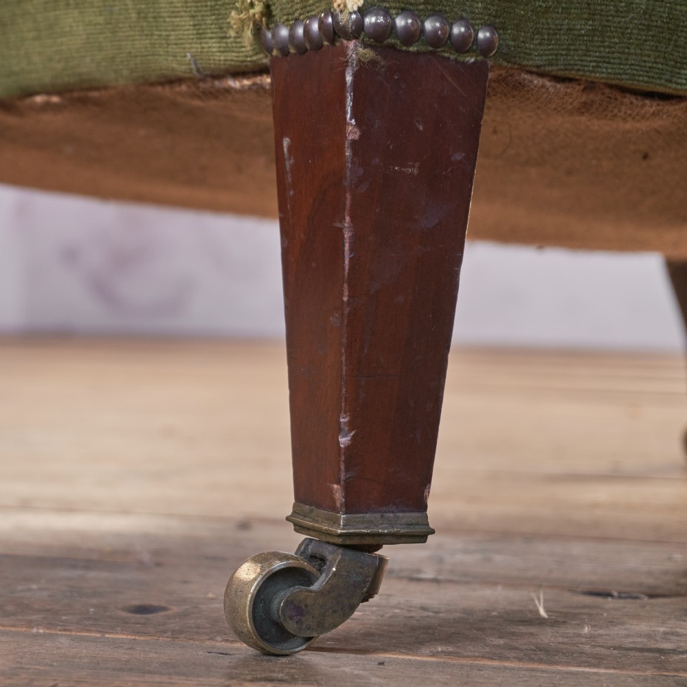 An antique green velvet wingback armchair featuring rolled arms, dark wooden legs, and brass castors. View VII