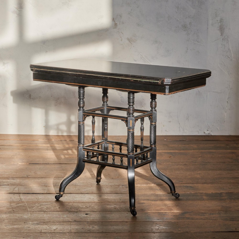 An ebonised late Victorian Aesthetic Movement card table with a rectangular fold-over top, incised gilt accents, spiral-turned legs, a lower spindle shelf, and brass castors.