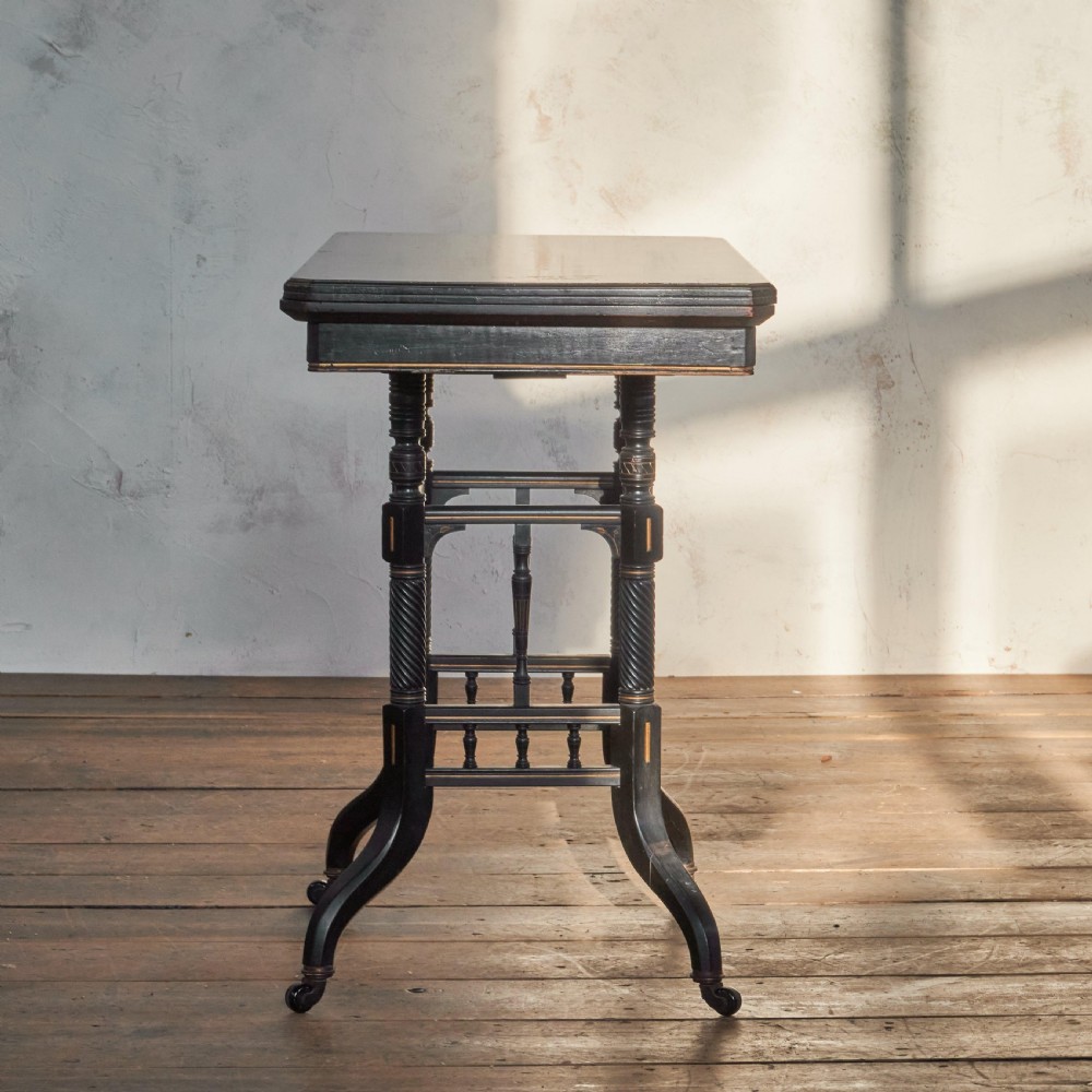 An ebonised late Victorian Aesthetic Movement card table with a rectangular fold-over top, incised gilt accents, spiral-turned legs, a lower spindle shelf, and brass castors. View II