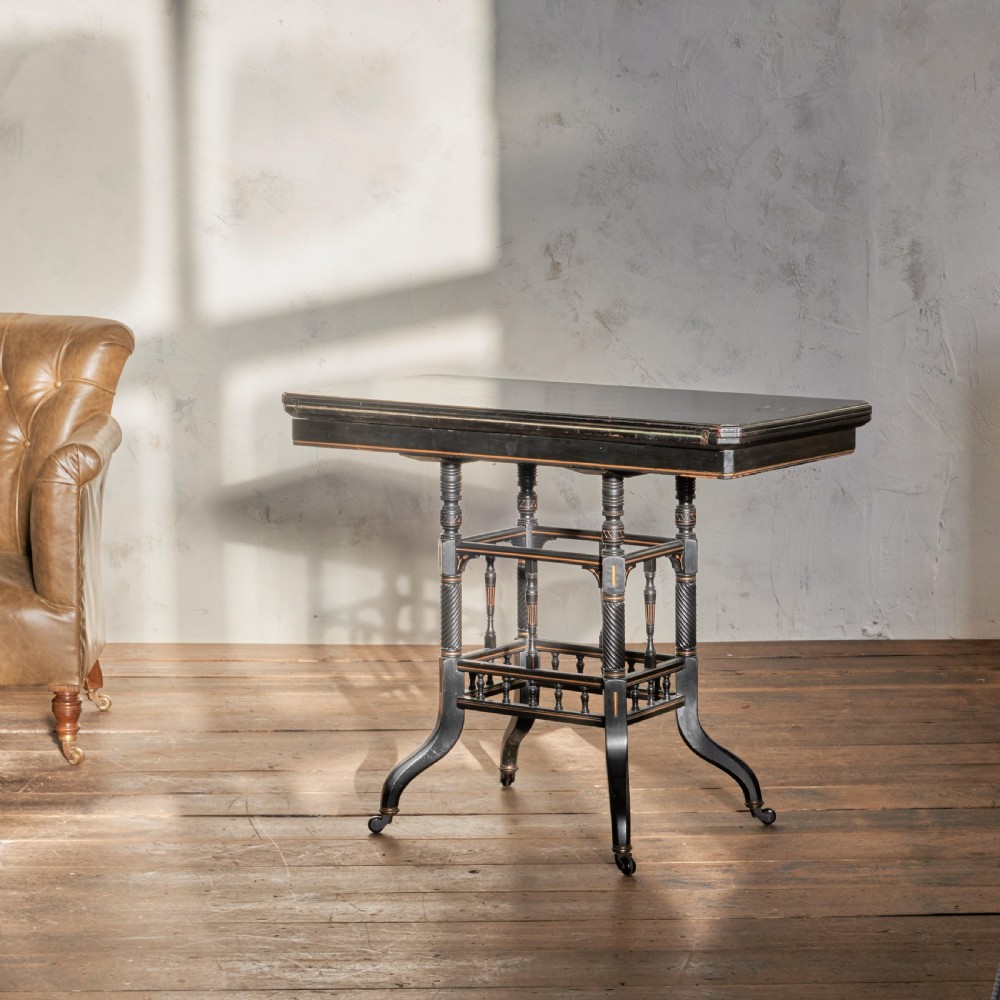 An ebonised late Victorian Aesthetic Movement card table with a rectangular fold-over top, incised gilt accents, spiral-turned legs, a lower spindle shelf, and brass castors. View V