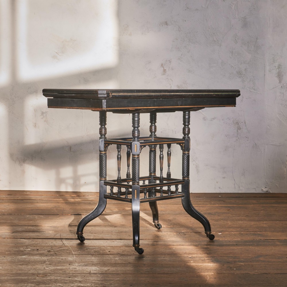 An ebonised late Victorian Aesthetic Movement card table with a rectangular fold-over top, incised gilt accents, spiral-turned legs, a lower spindle shelf, and brass castors. View VI