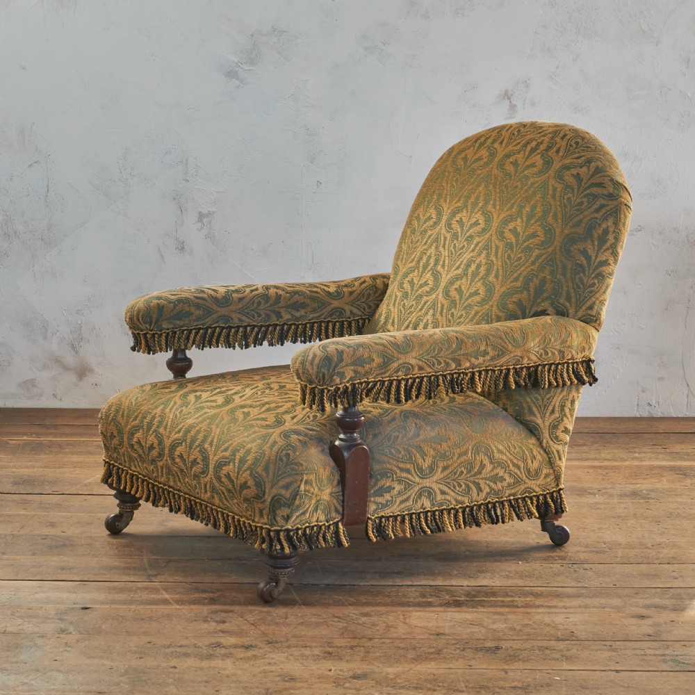 A Victorian open armchair upholstered in a patterned green and gold fabric with fringe trim on turned legs with brass castors.