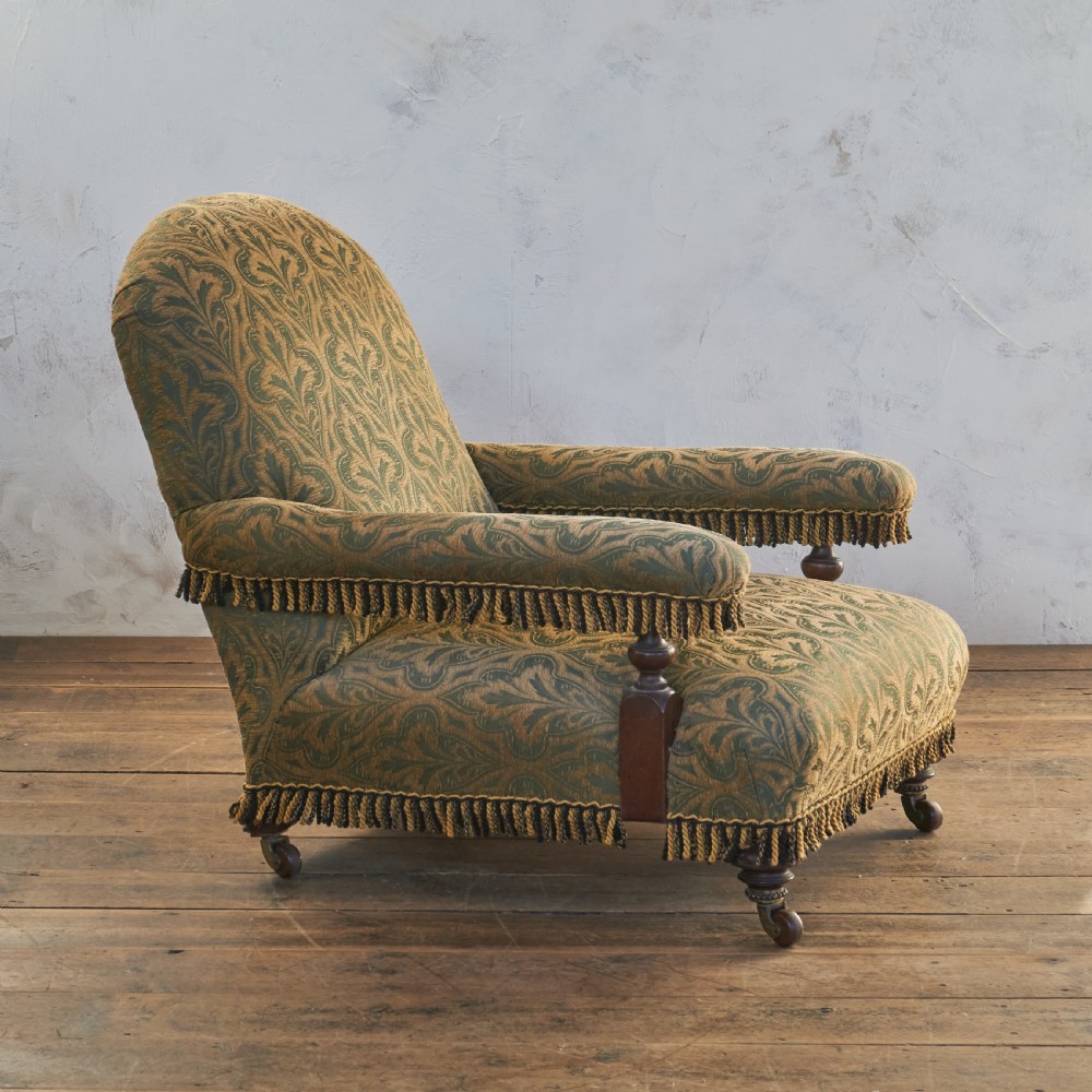 A Victorian open armchair upholstered in a patterned green and gold fabric with fringe trim on turned legs with brass castors. View II