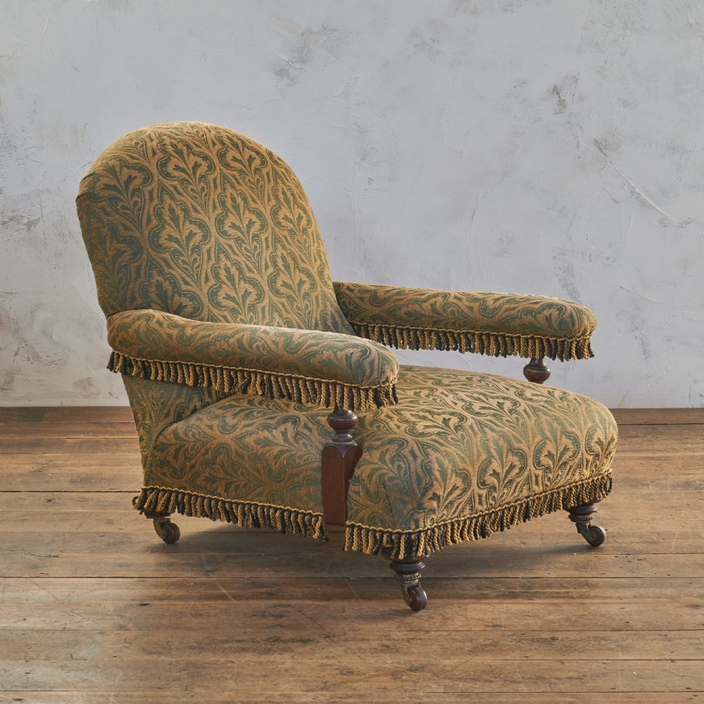 A Victorian open armchair upholstered in a patterned green and gold fabric with fringe trim on turned legs with brass castors. View III