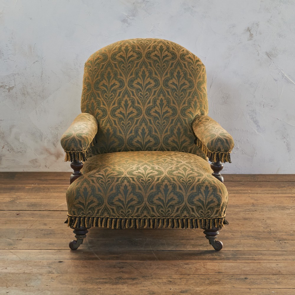 A Victorian open armchair upholstered in a patterned green and gold fabric with fringe trim on turned legs with brass castors. View IV