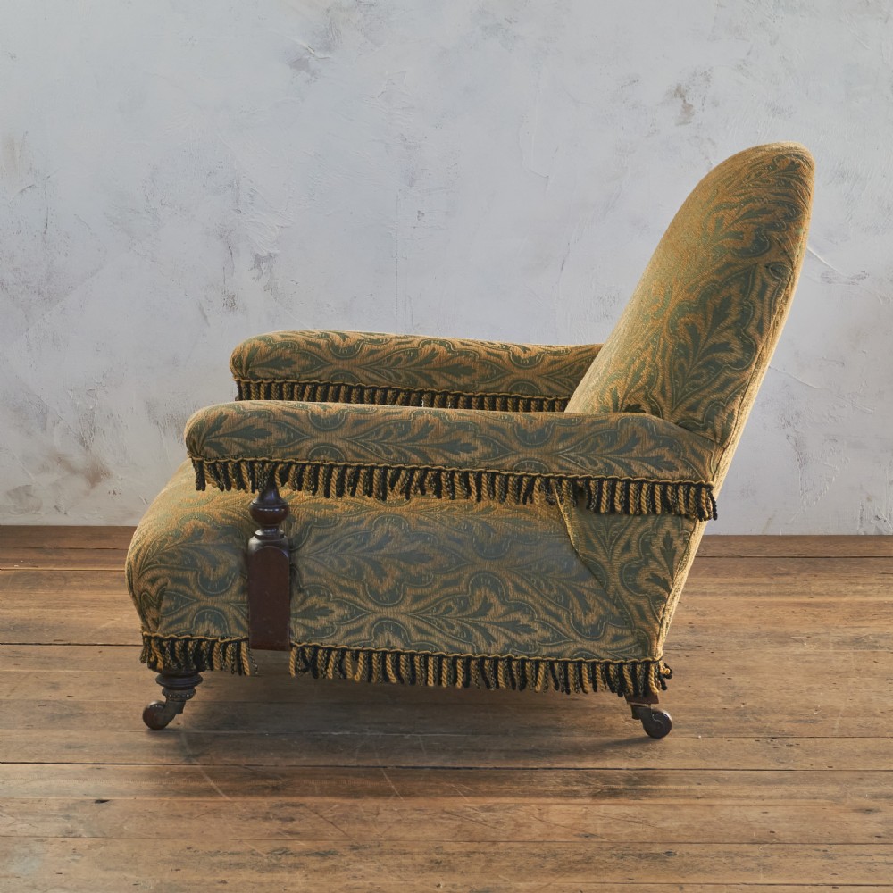 A Victorian open armchair upholstered in a patterned green and gold fabric with fringe trim on turned legs with brass castors. View V