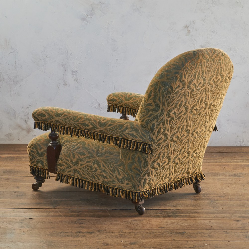 A Victorian open armchair upholstered in a patterned green and gold fabric with fringe trim on turned legs with brass castors. View VI
