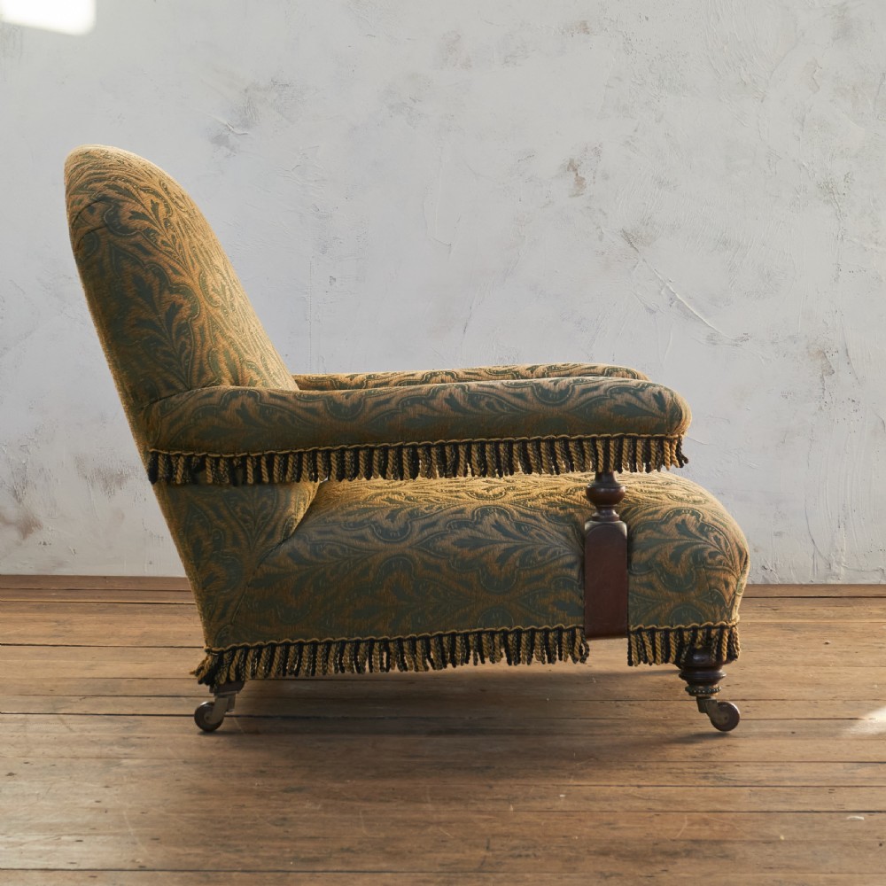 A Victorian open armchair upholstered in a patterned green and gold fabric with fringe trim on turned legs with brass castors. View VIII