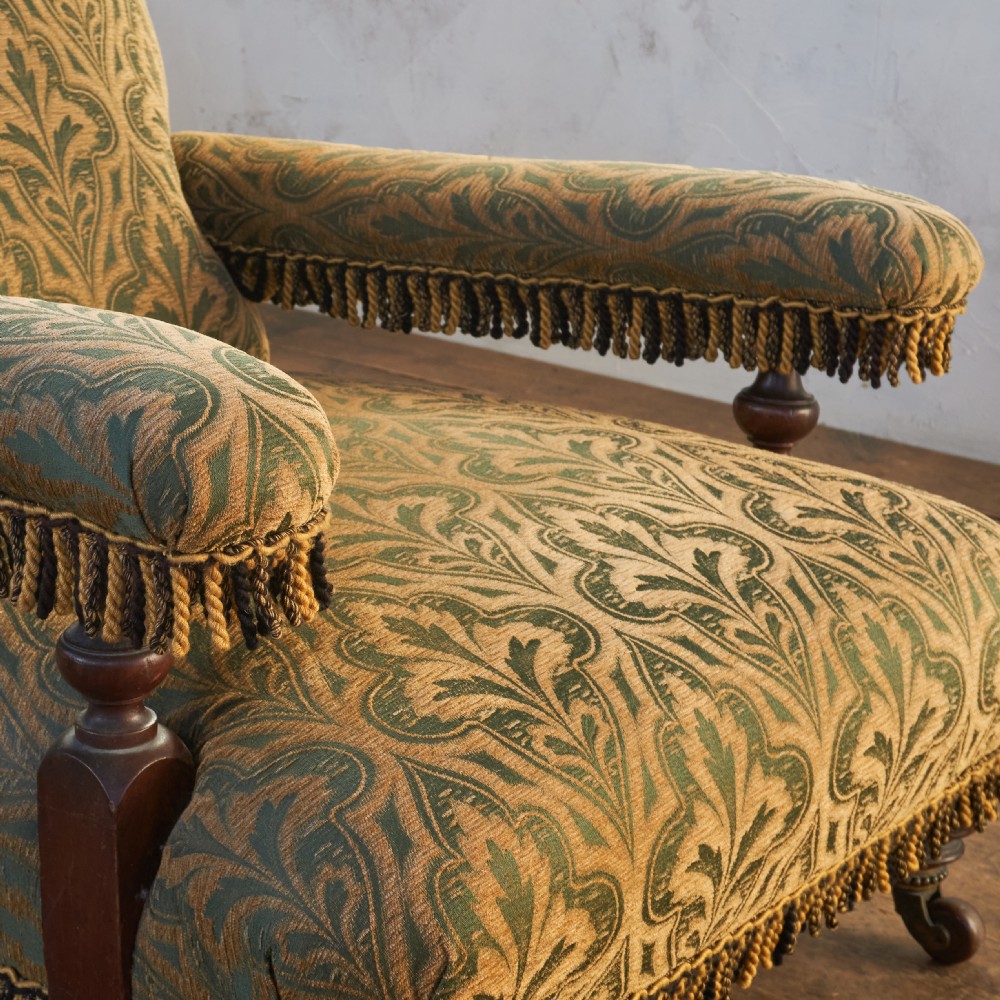 A Victorian open armchair upholstered in a patterned green and gold fabric with fringe trim on turned legs with brass castors. View IX
