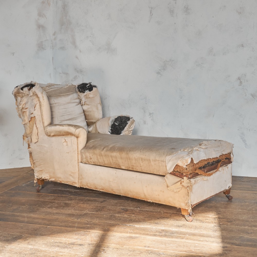 A vintage upholstered chaise longue with exposed wooden legs on castors and worn fabric revealing internal stuffing.