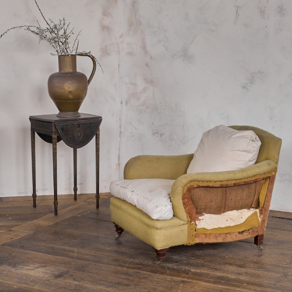 An antique deep-seated armchair with exposed hessian framing, olive green fabric, and turned wooden legs with castors. View V