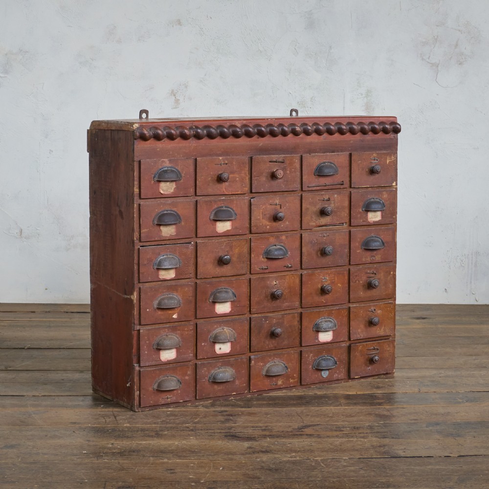 An antique multi-drawer wooden cabinet featuring a red painted finish, numerous small drawers with metal cup handles, and decorative wooden spool moulding along the top edge. View II