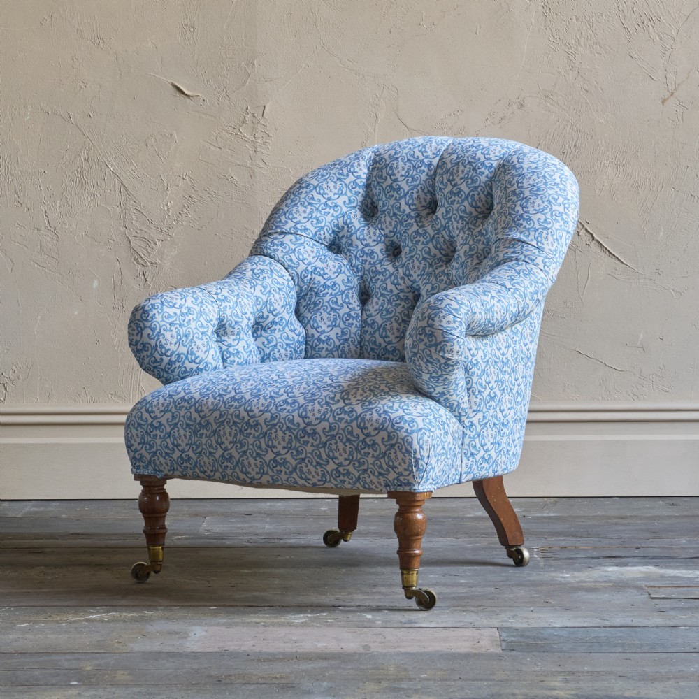 An antique tub armchair upholstered in a blue and white patterned fabric, featuring deep buttoning, turned front legs, and brass castors.