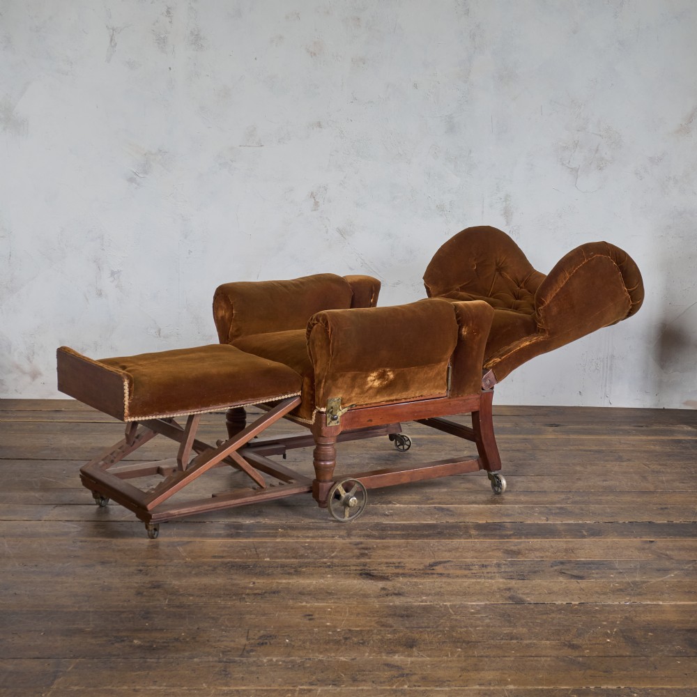 An antique Victorian mechanical reclining armchair upholstered in brown velvet with a button-tufted backrest, padded armrests, and an extendable footrest on brass castors. View V